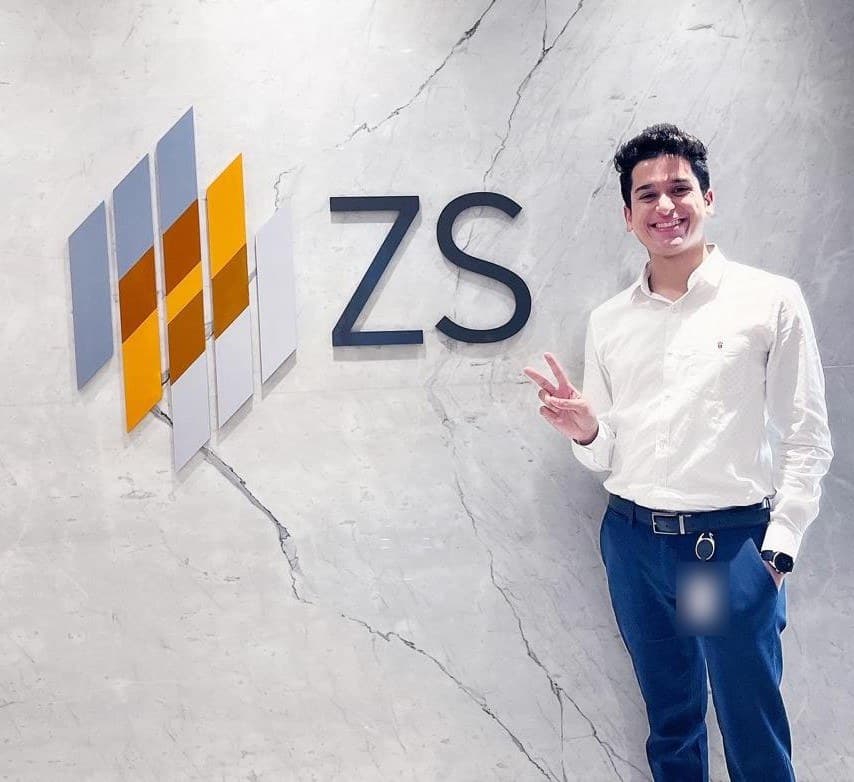 Kushagra standing beside the ZS logo on an office wall.